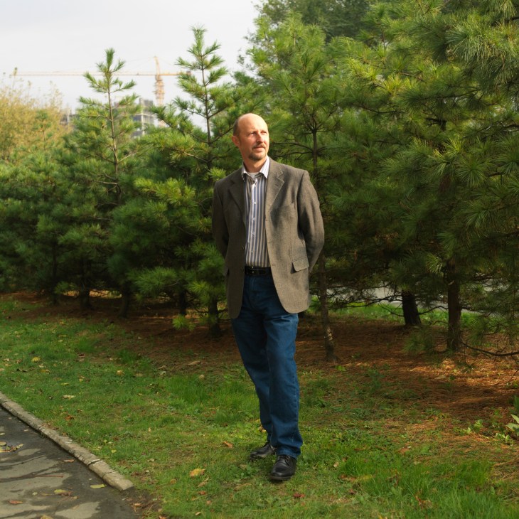 Andrei Kalachinsky standing in a park with evergreen trees and a church visible in the background.