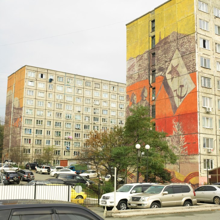 Soviet-era apartment buildings with large murals featuring communist symbols and red propaganda imagery.