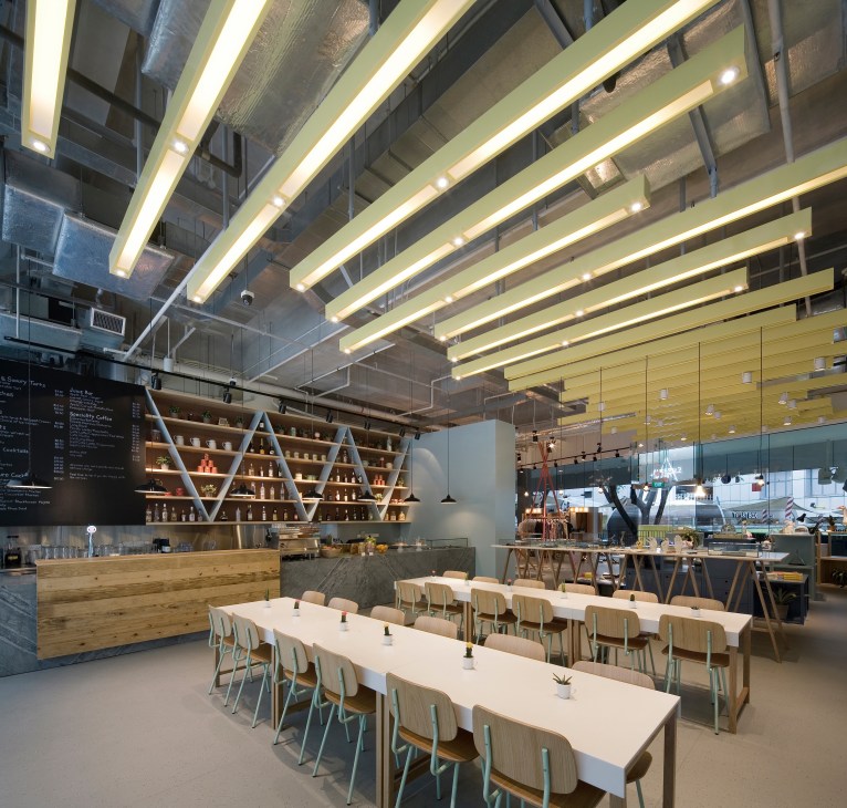 Modern restaurant interior with long white tables, wooden chairs, geometric shelving, and linear ceiling lights.