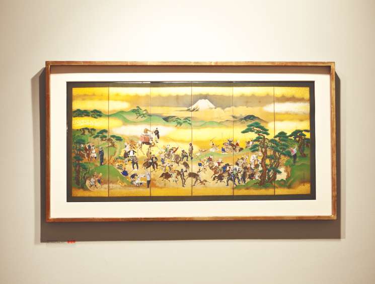 Traditional Japanese six-panel folding screen artwork depicting a crowded military scene with Mount Fuji in background, displayed in a wooden frame on gallery wall.