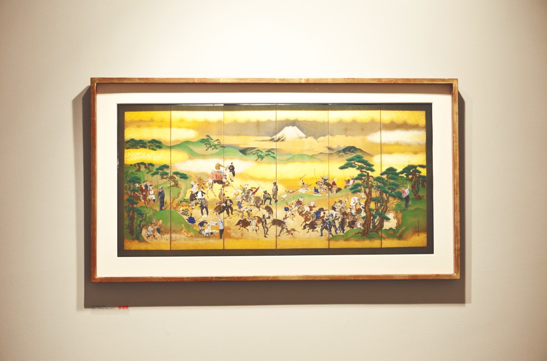 Traditional Japanese six-panel folding screen artwork depicting a crowded military scene with Mount Fuji in background, displayed in a wooden frame on gallery wall.
