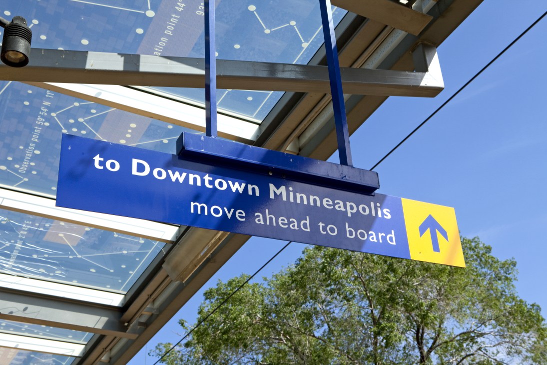 Blue directional sign reading "to Downtown Minneapolis move ahead to board" with yellow arrow under pergola structure.