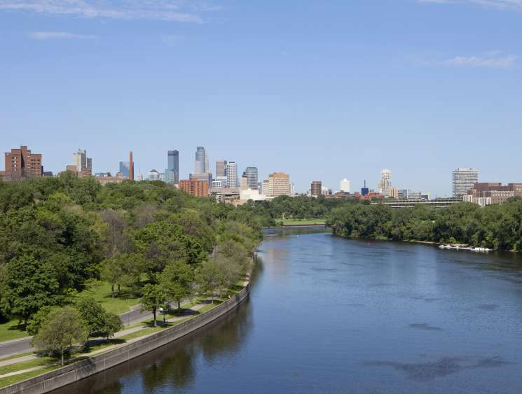 Minneapolis skyline with modern glass and metal buildings along the Mississippi River surrounded by green trees.