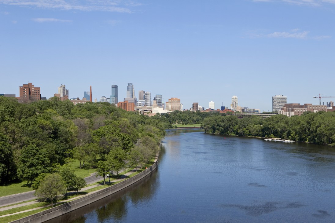 Minneapolis skyline with modern glass and metal buildings along the Mississippi River surrounded by green trees.