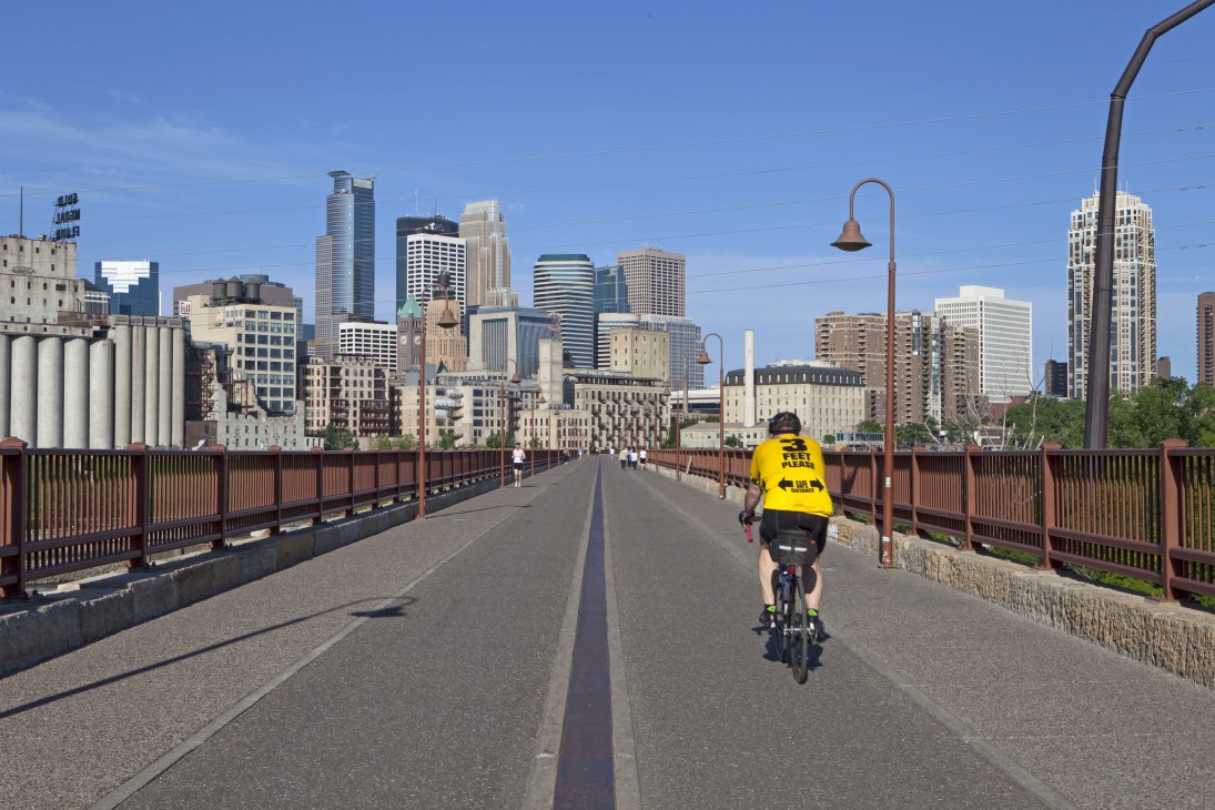 Cyclist in yellow vest riding across Stone Arch Bridge with Minneapolis skyline in background.