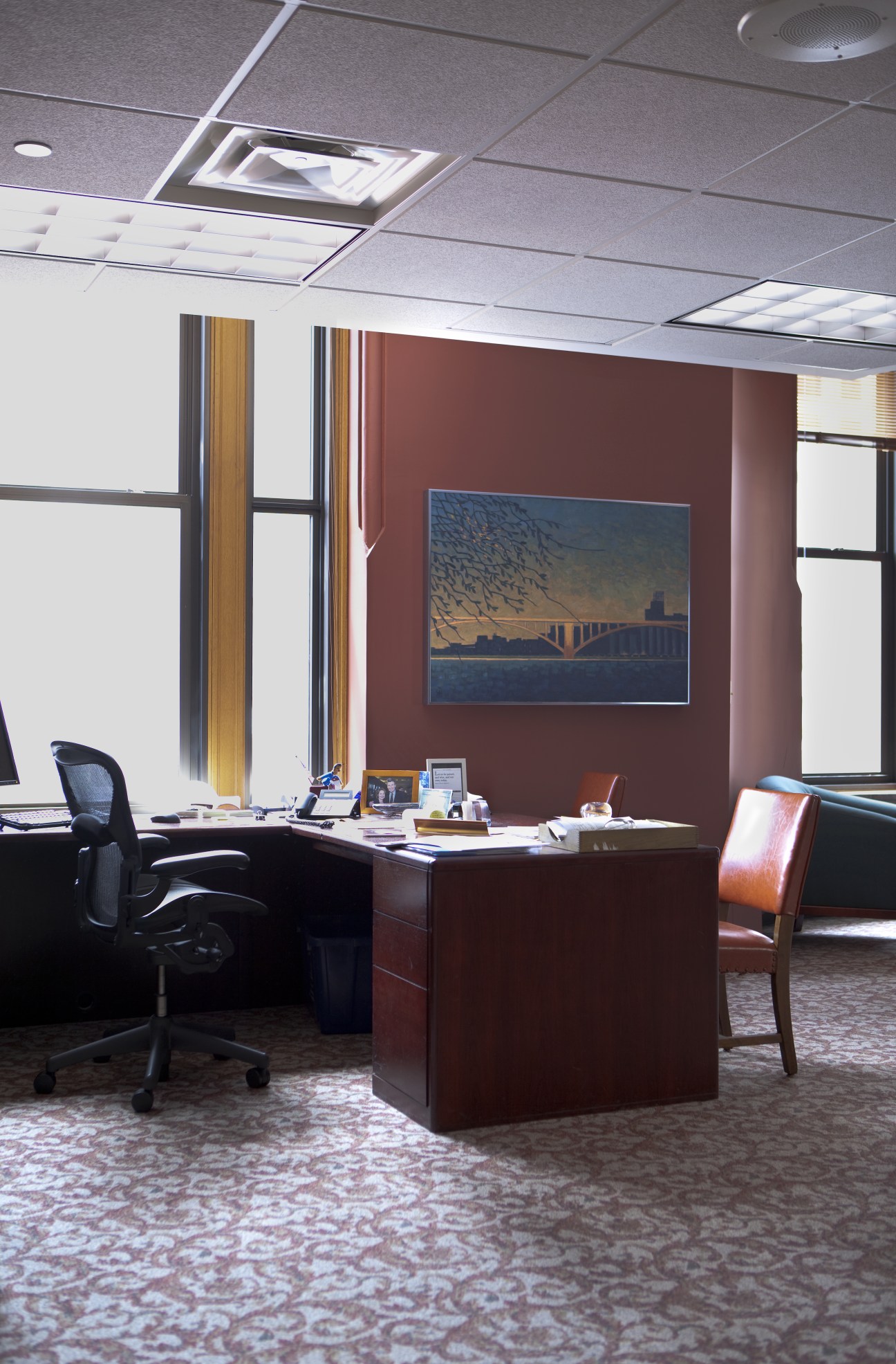 Mayor RT Rybak's office desk with burgundy wall, windows, and artwork of a bridge.