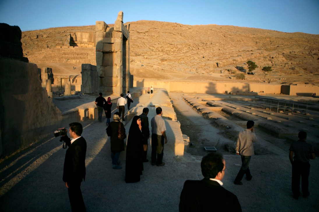 Tourists visit ancient Persian ruins at Persepolis with stone columns and desert landscape.