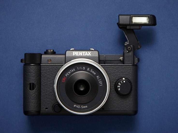 Pentax Q compact camera with interchangeable lens and built-in flash on blue background.