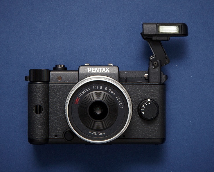 Pentax Q compact camera with interchangeable lens and built-in flash on blue background.