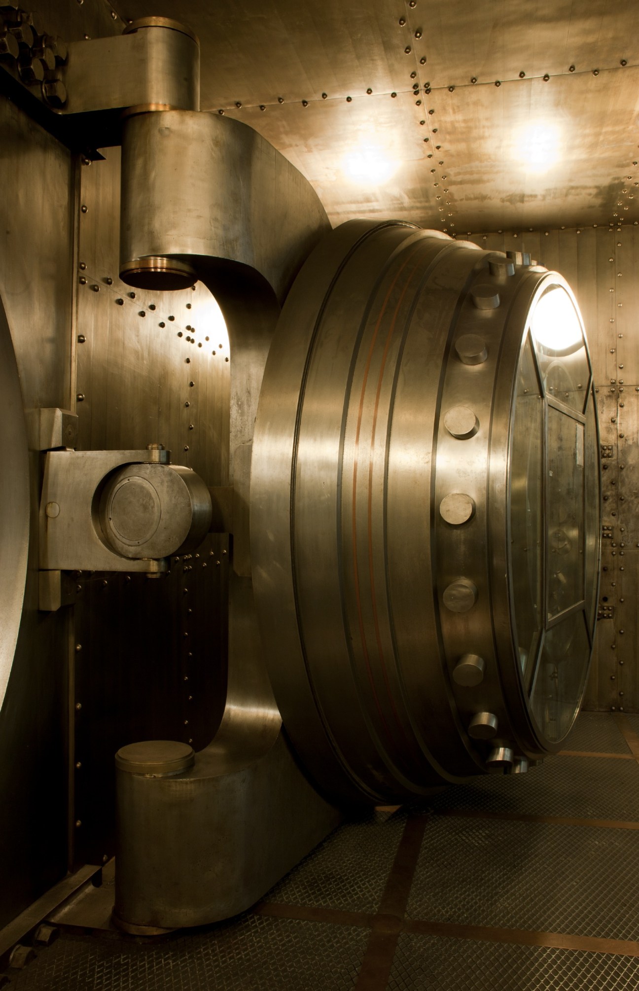Large circular steel bank vault door with riveted metal plating and mechanical locking mechanism.