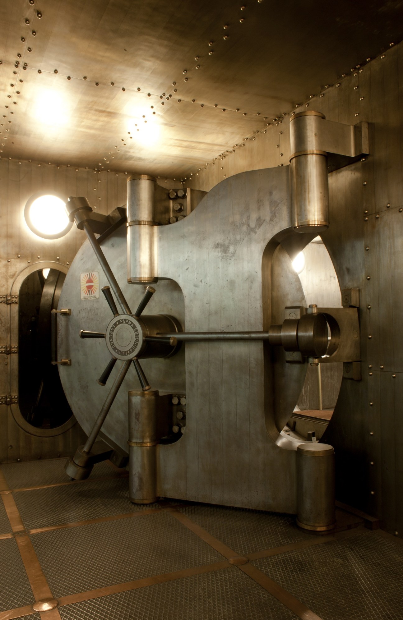 Interior of a bank vault with steel door, combination lock, and riveted walls illuminated by overhead lights.