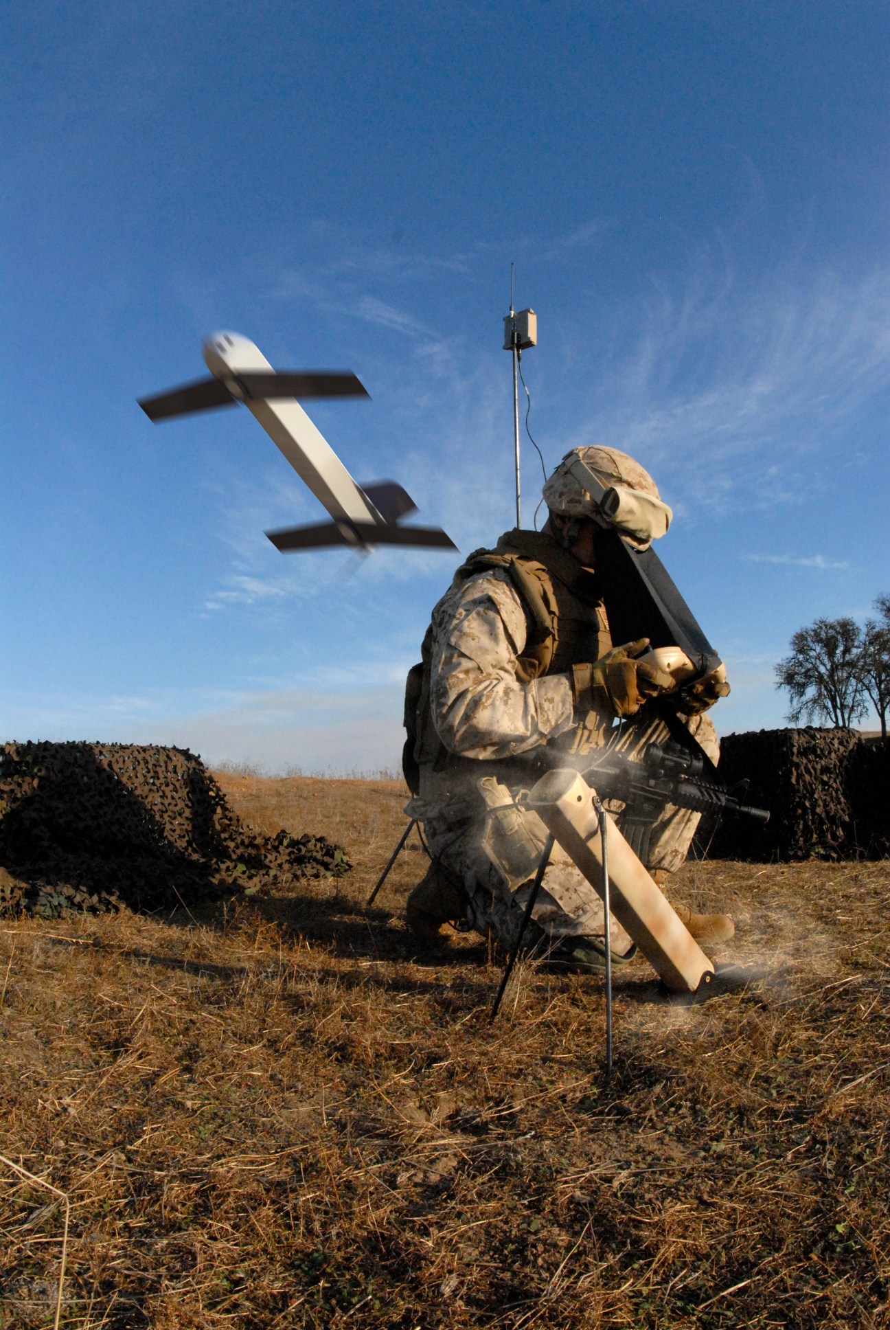 Soldier operating a Switchblade drone in an arid field with the unmanned aircraft visible in flight above.