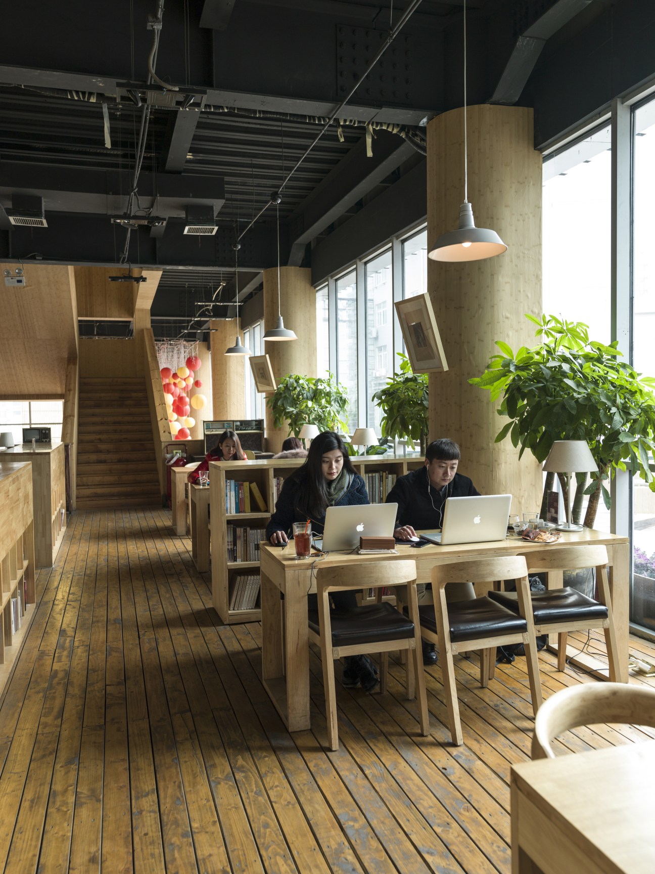 Two people work on laptops at a wooden table in a modern café library with exposed ceiling and large windows.
