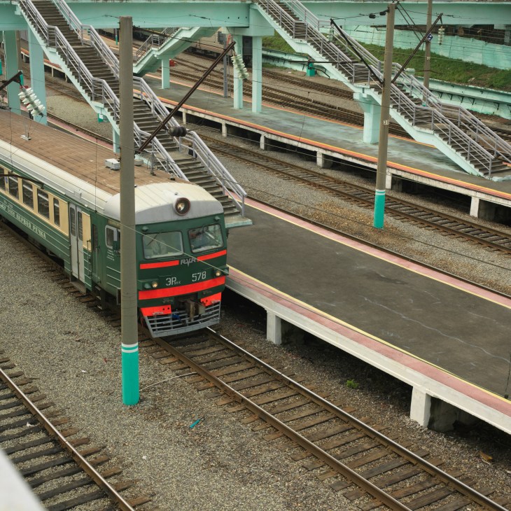A green and red local train at Vladivostok station with platforms and overhead structures.