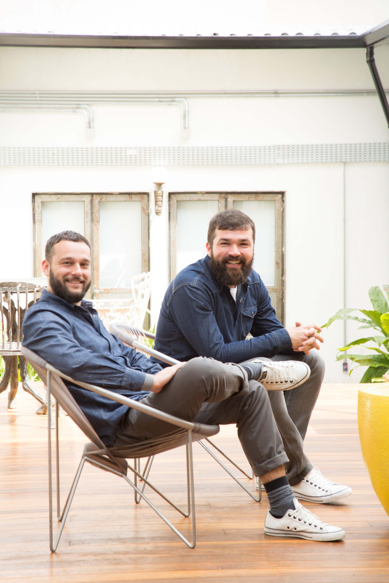 Two men sitting on a wooden deck outside a white building with glass doors.