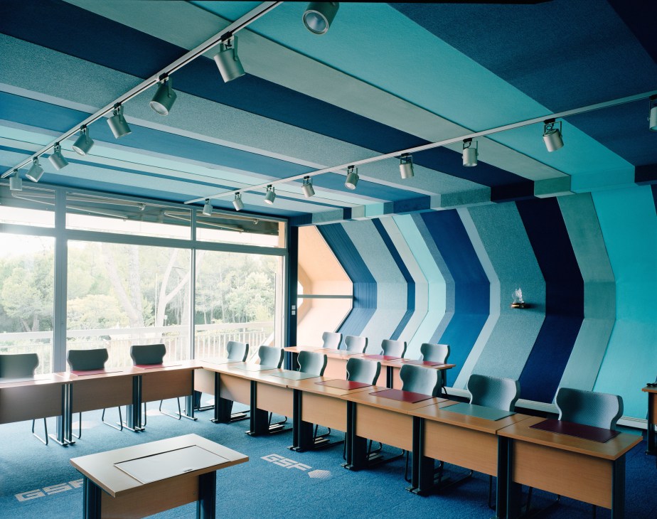 Modern office meeting room with turquoise striped wall panels, wooden desks, pale blue chairs, track lighting, and forest views through large windows.
