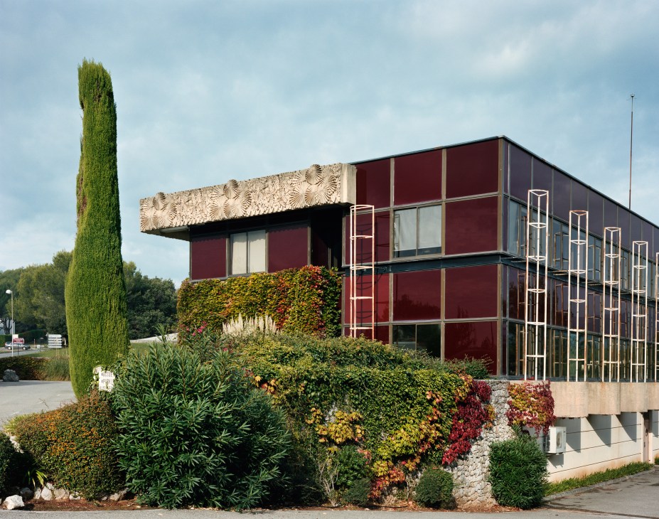 Modern physics lab building with burgundy panels and decorative facade surrounded by colorful flowering shrubs and tall cypress trees.