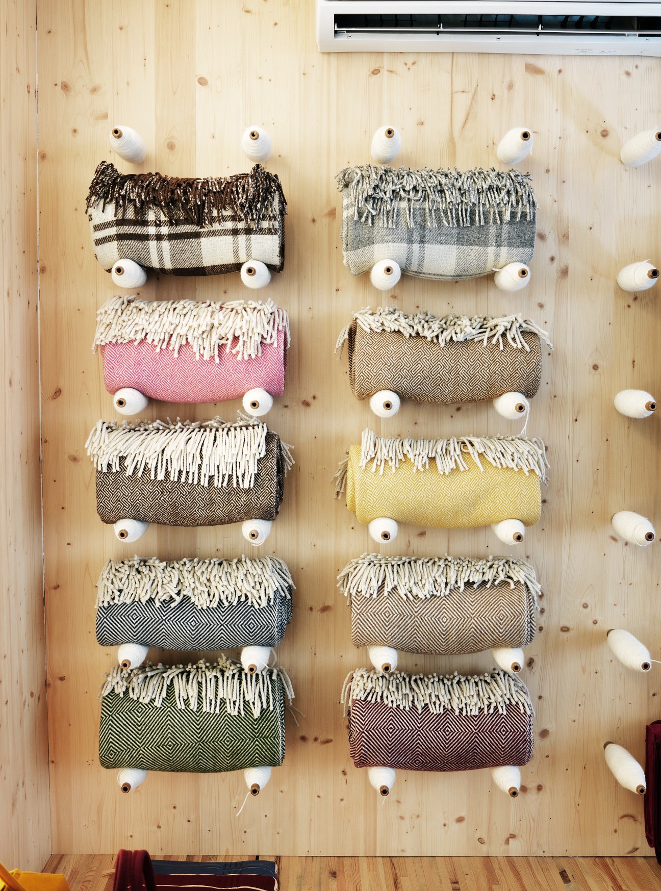 Rolled Portuguese wool blankets and throws with fringed edges displayed on a light wood wall with white pegs.