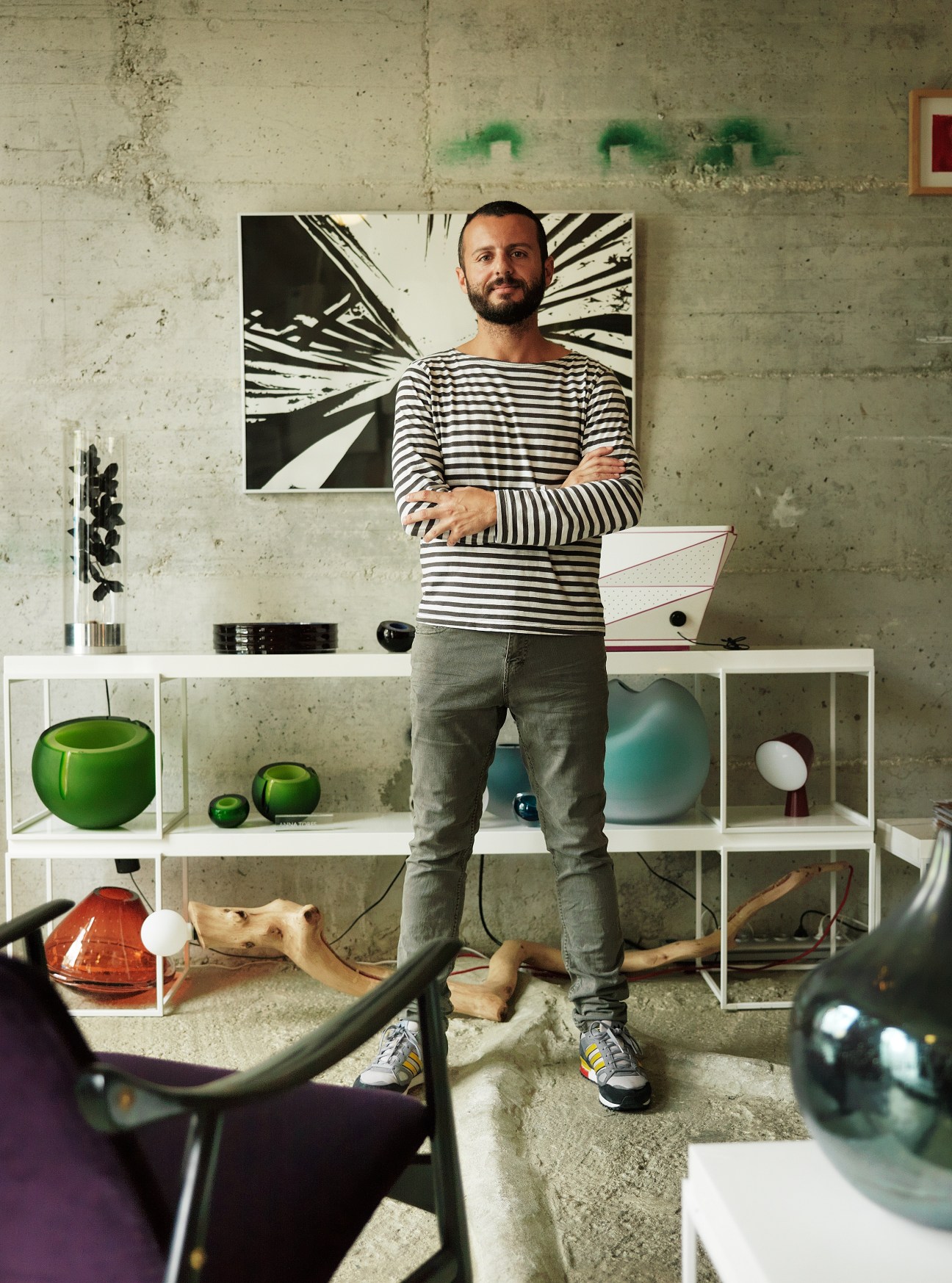 Man in striped shirt standing in front of shelving unit displaying design objects and artwork.