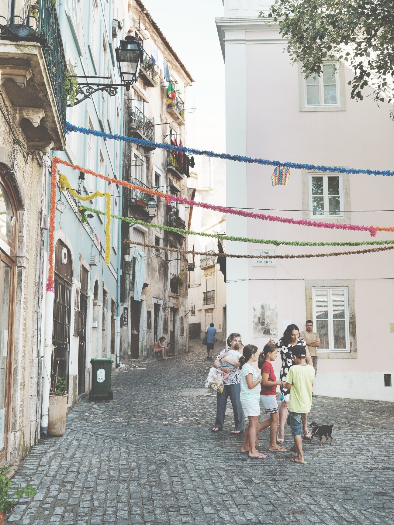 Colorful decorative bunting strung across a narrow cobblestone street in Lisbon's Mouraria neighbourhood with historic buildings and local residents.