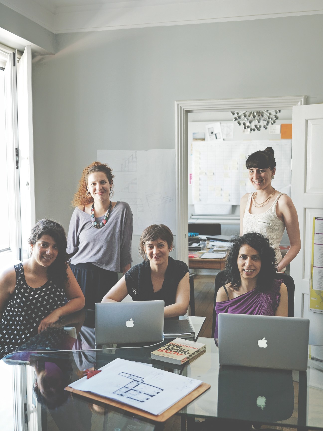 Five women pose together in a bright office space with laptops, architectural drawings and design materials visible.
