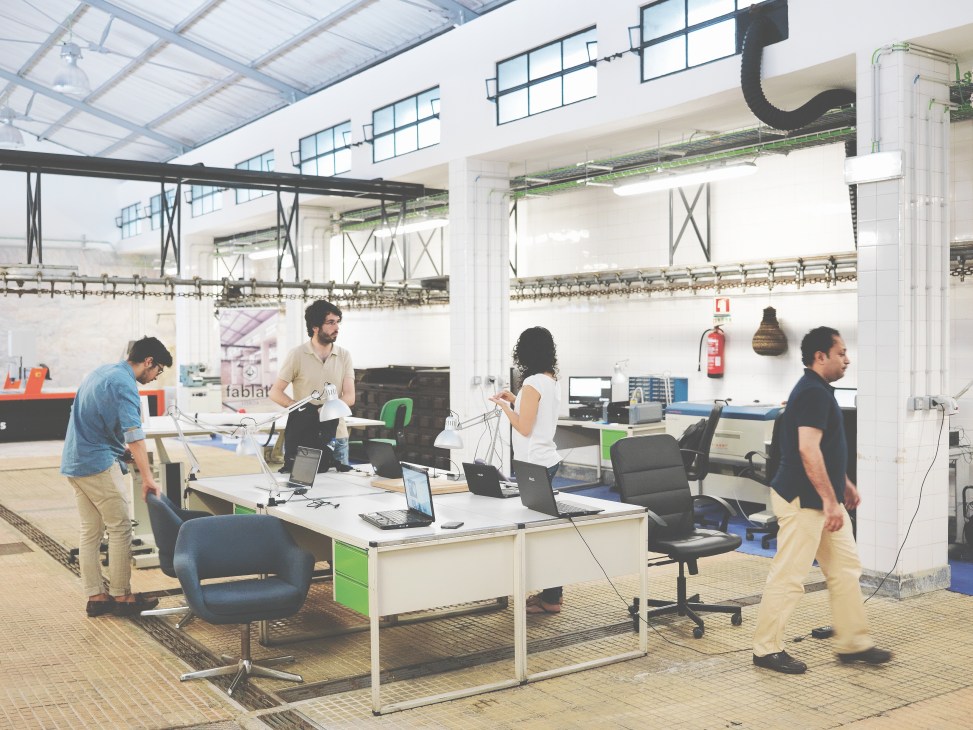 Workers at desks inside FabLab Lisboa, a bright industrial space with skylights and modern equipment.