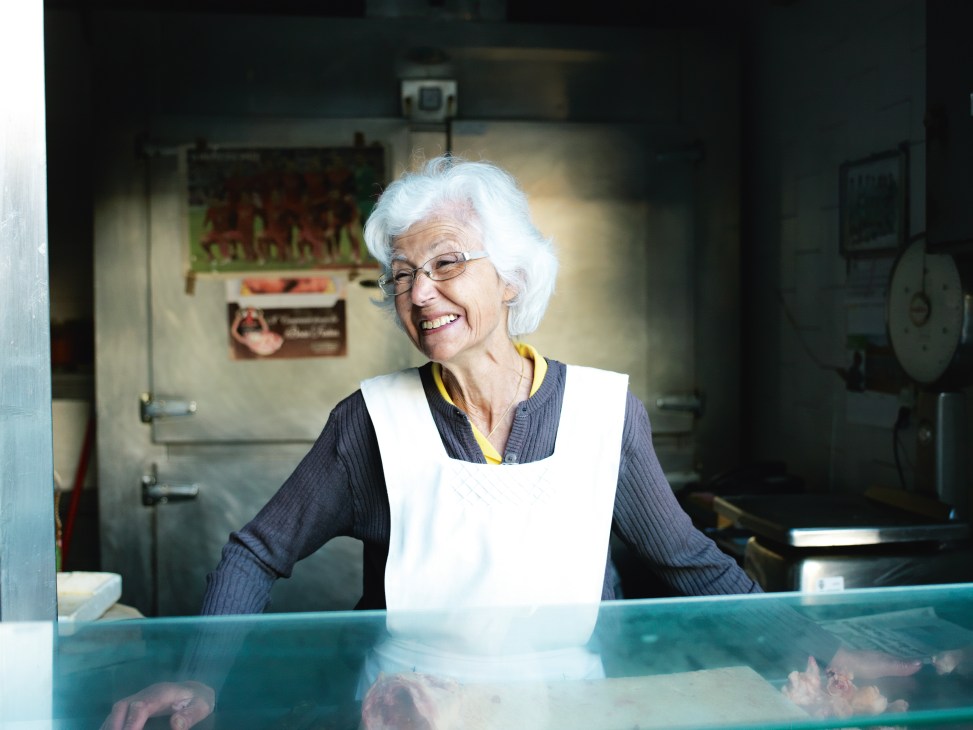 Marilia Brandão, a 73-year-old vendor with white hair and glasses, smiles behind a market counter at Mercado do Bolhão.
