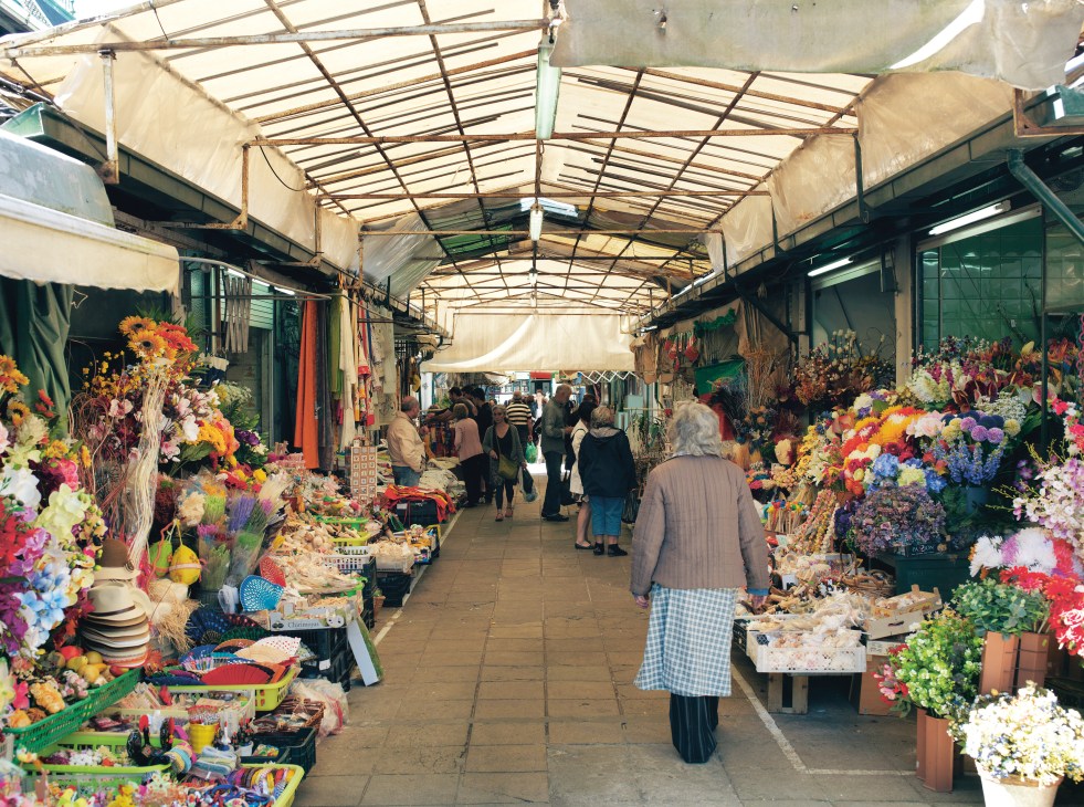 Covered flower market corridor with colorful blooms on both sides and shoppers walking through.