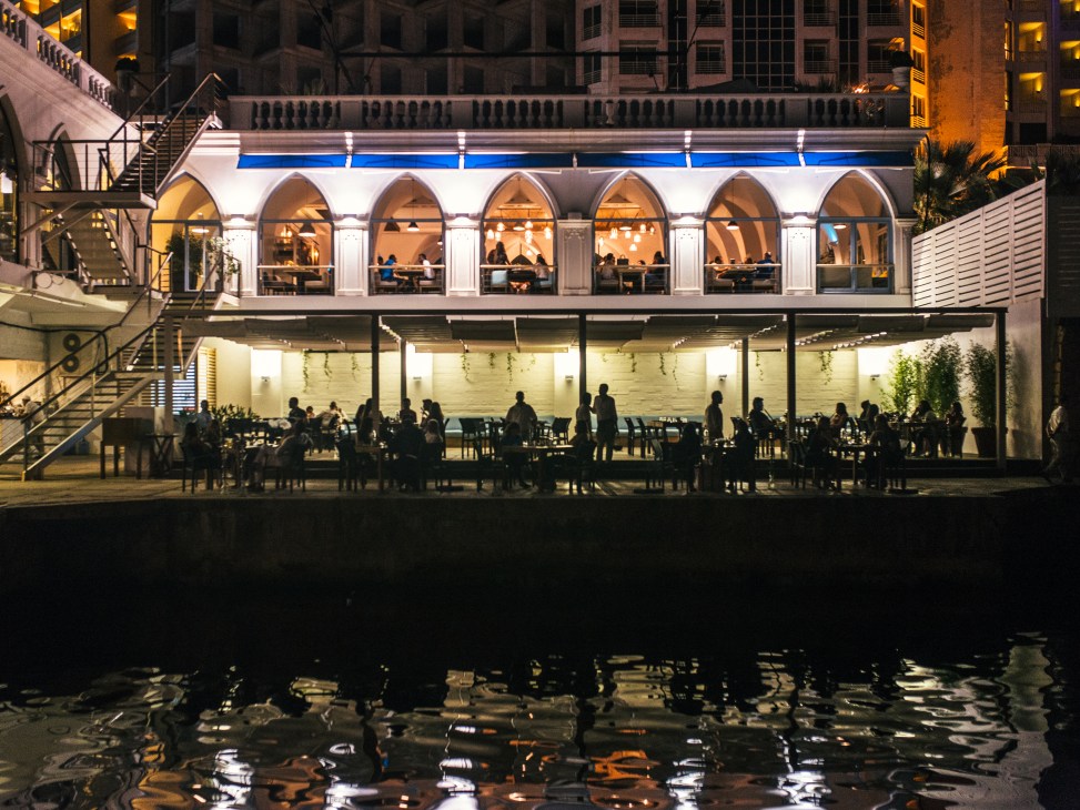 Illuminated waterfront restaurant with arched windows and diners visible on two levels at night.
