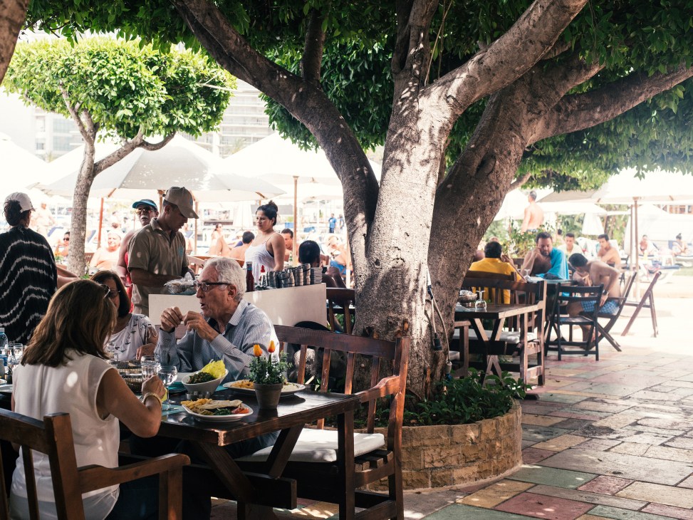 Diners enjoying lunch at tables under large trees at a beachfront restaurant in Beirut.
