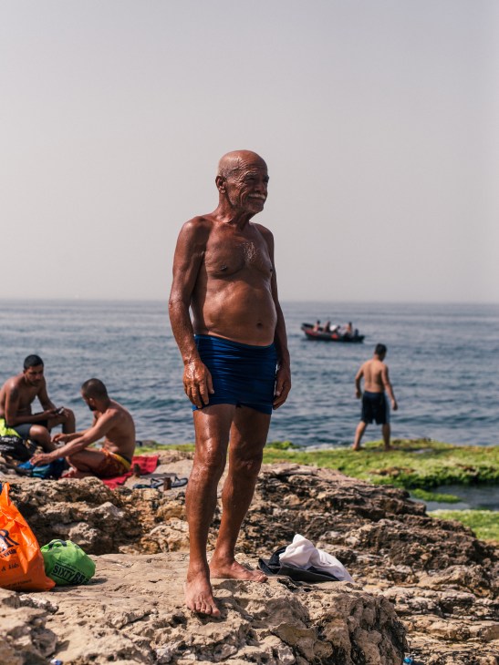 An older man in blue shorts stands on rocky Beirut coastline with other swimmers and boats visible in the background.