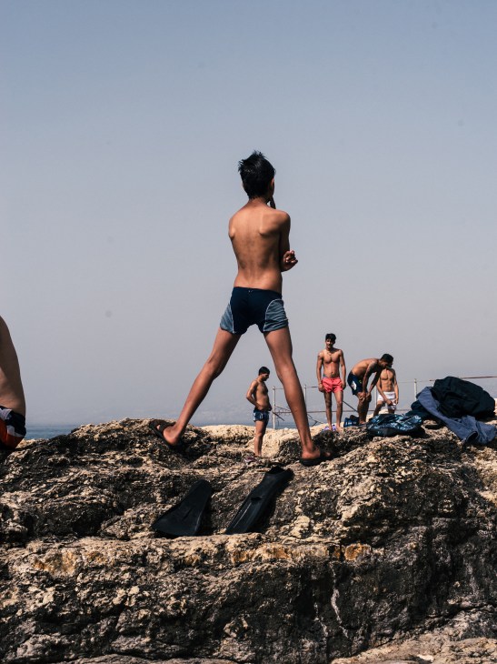 Young men in swimming shorts posing on rocky beach with clear blue sky.