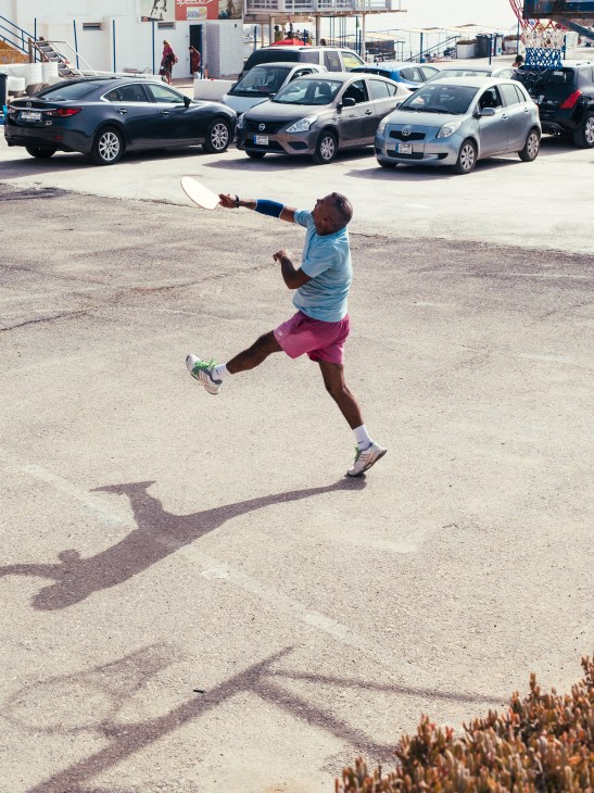 Man jumping to catch a frisbee in a parking lot with cars parked behind him.