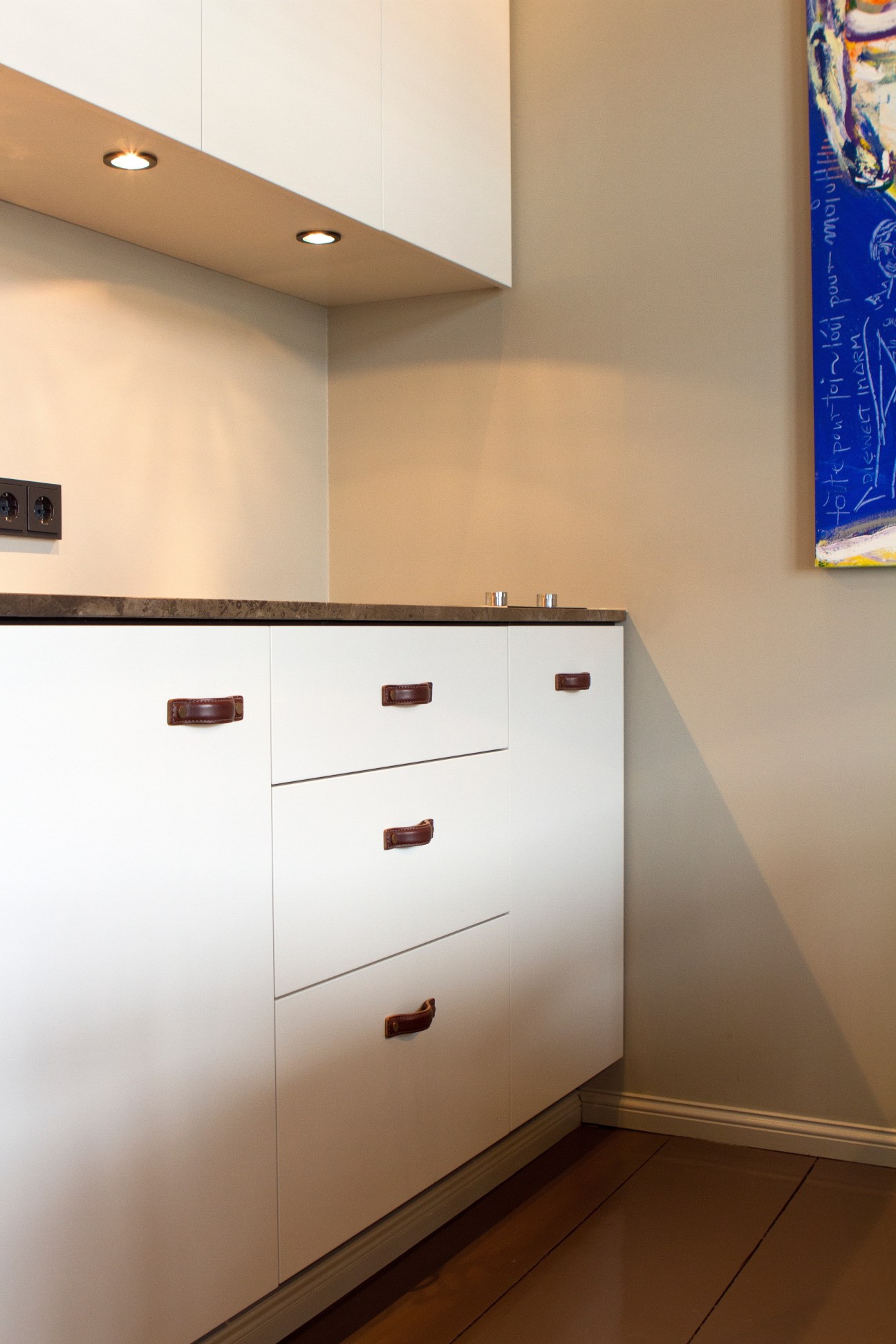 Minimalist kitchen with white cabinetry, leather handles, recessed lighting, and beige walls.