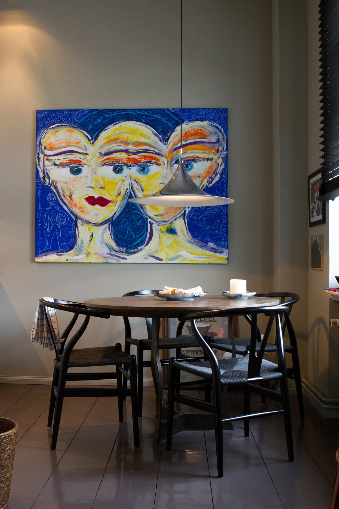 Modern dining room with round wooden table, black chairs, pendant light, and colorful abstract portrait artwork on blue background.