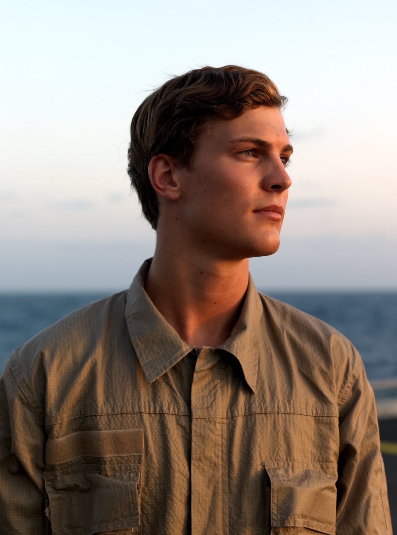 A young man in a tan military shirt looks toward the horizon while standing on a ship at sea.