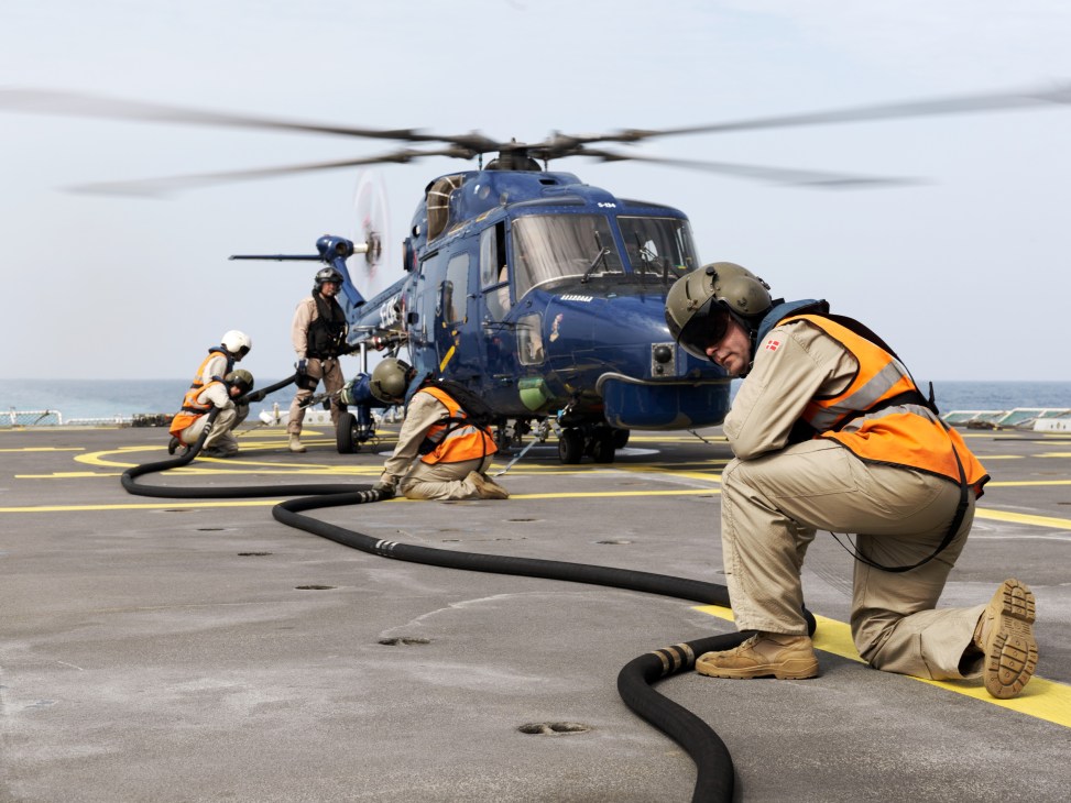 Helicopter launch team members in orange vests refuel a blue military helicopter on the ship's deck.