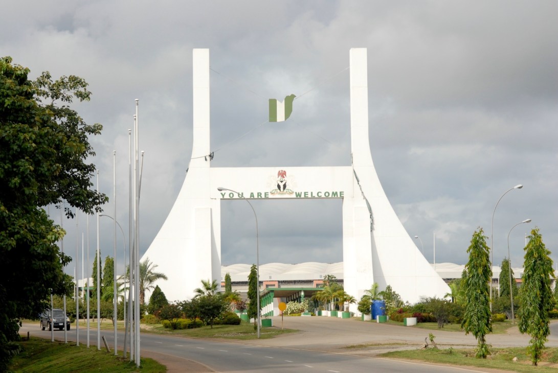 White monumental gate structure with 'YOU ARE WELCOME' text and Nigerian emblem, flanked by tall poles and landscaping.