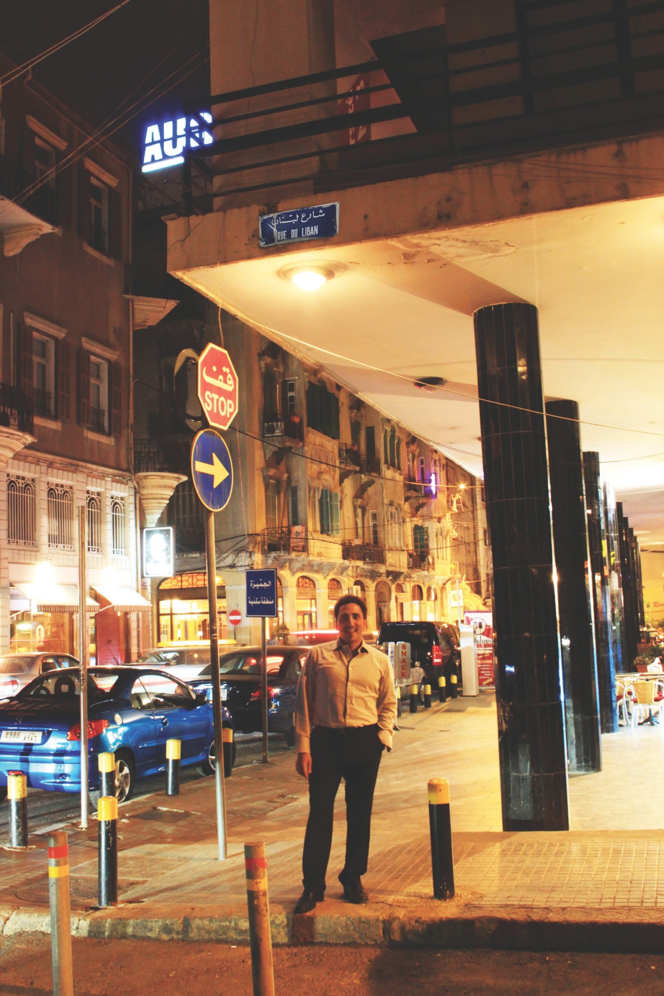 Man standing on a Beirut street at night near traffic signs and parked cars with historic buildings.