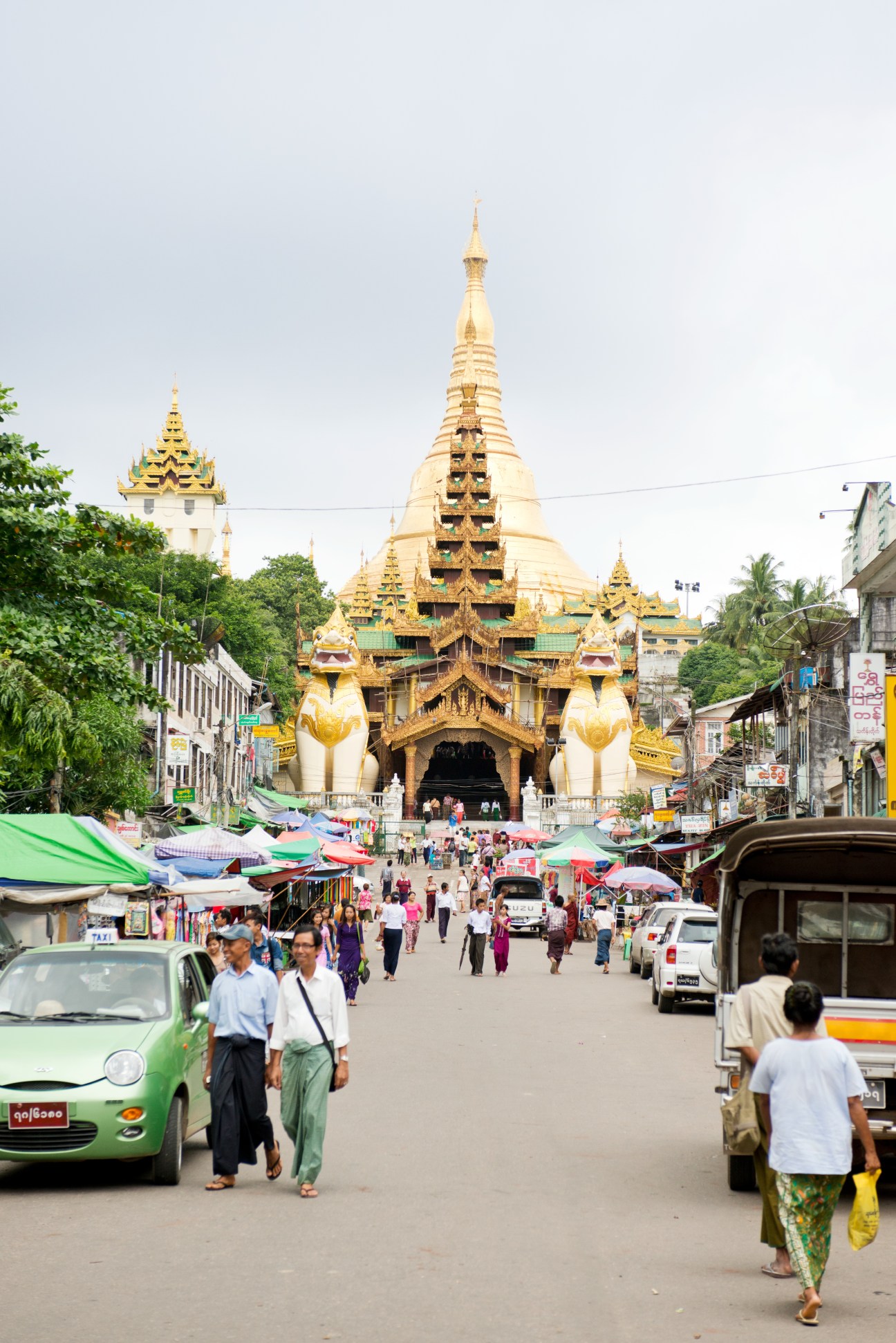 Shwedagon Pagoda's golden spires rise above a busy street market in Yangon filled with pedestrians, colorful umbrellas and vehicles.