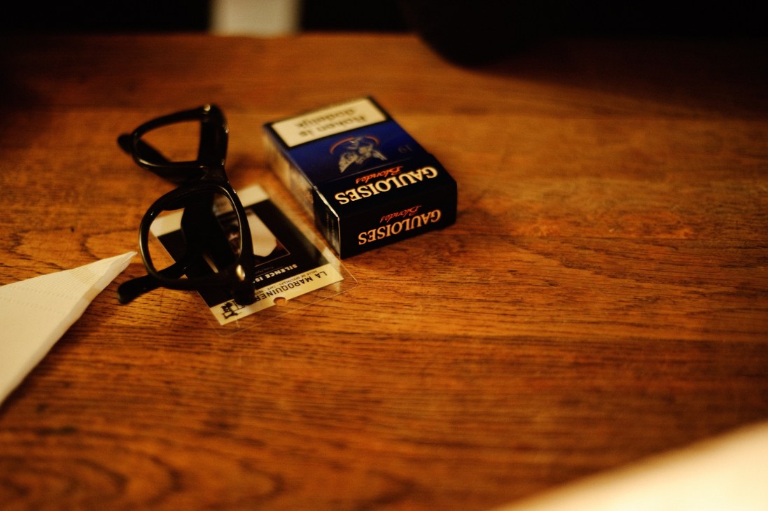 Glasses, cigarette pack, and backstage pass arranged on a wooden surface.