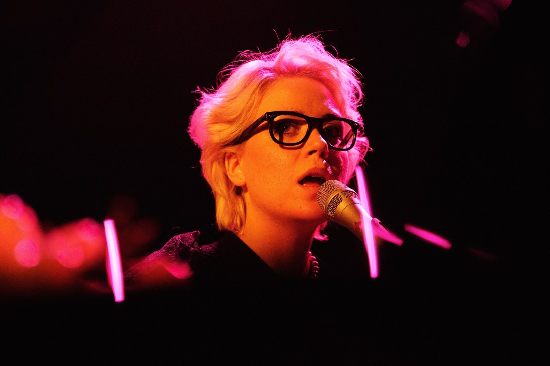 Swedish singer-songwriter Frida Hyvönen performing at microphone wearing glasses under pink and orange stage lighting.