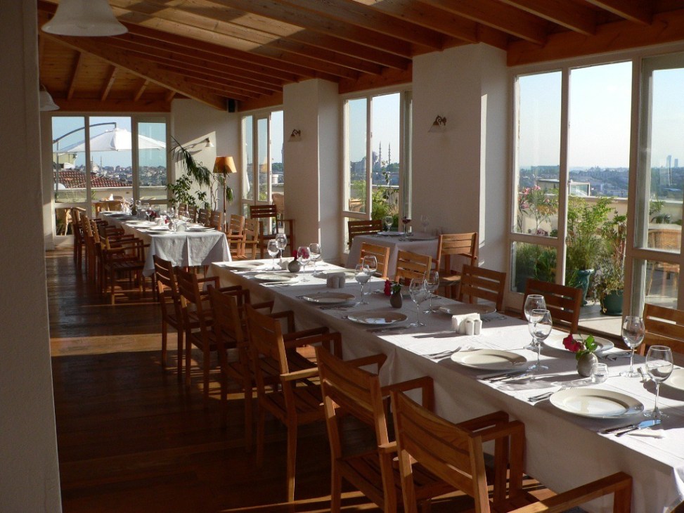 Bright dining room with wooden beams, set tables, and expansive windows overlooking a city skyline.