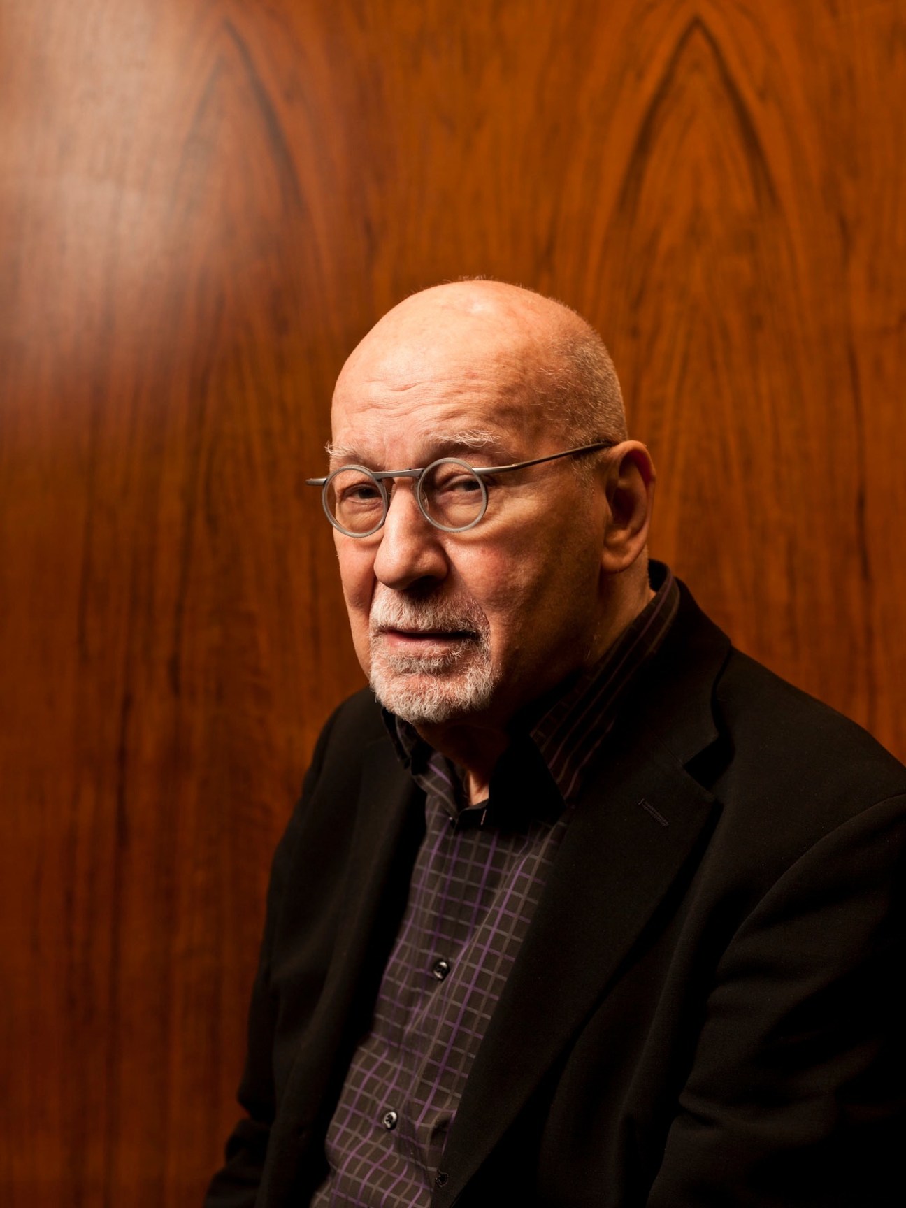 Portrait of George Lois, an older man wearing glasses and a black blazer against a wood-paneled background.
