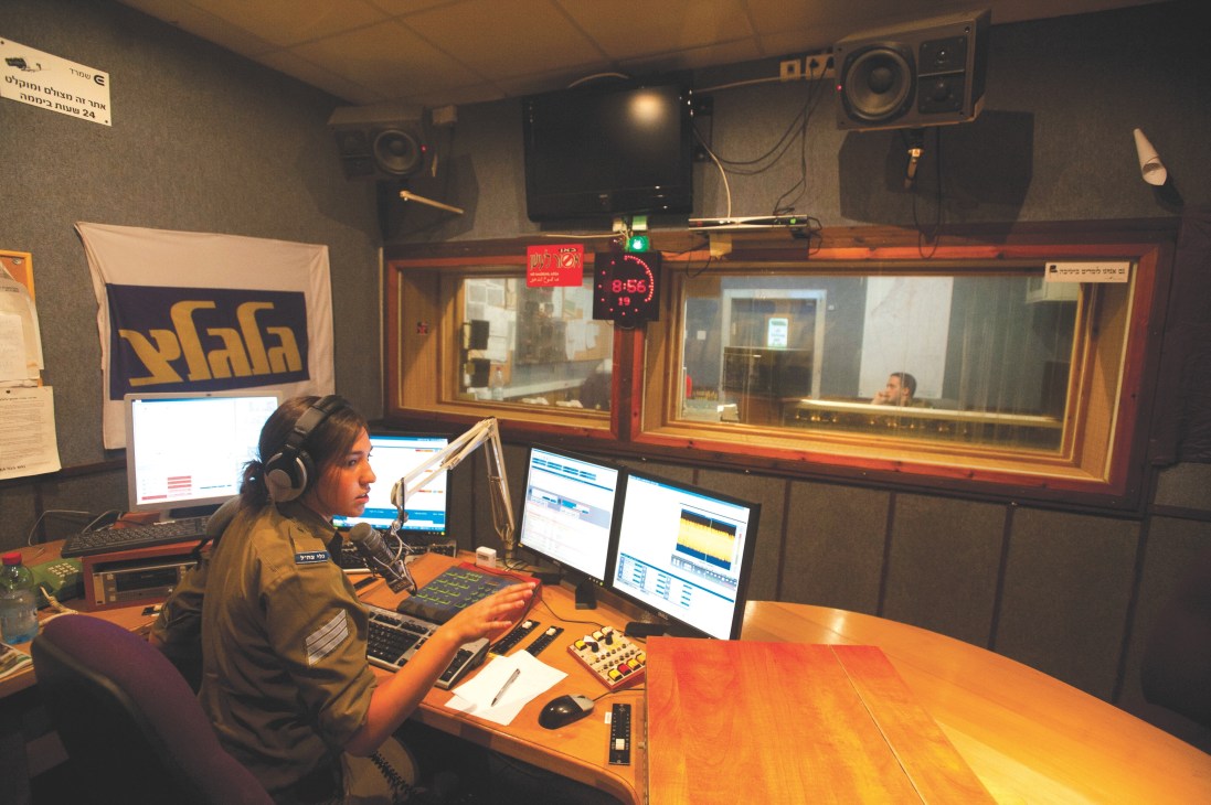 Israeli soldier Erga Shiloni in khaki uniform operating broadcast equipment at Galgalatz radio station studio desk.