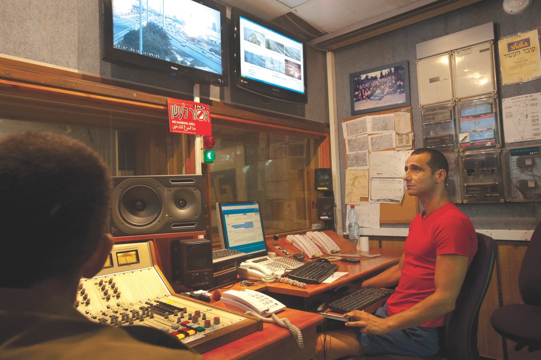 Man in red shirt sits at radio studio control desk with mixer, computer, and monitors visible.