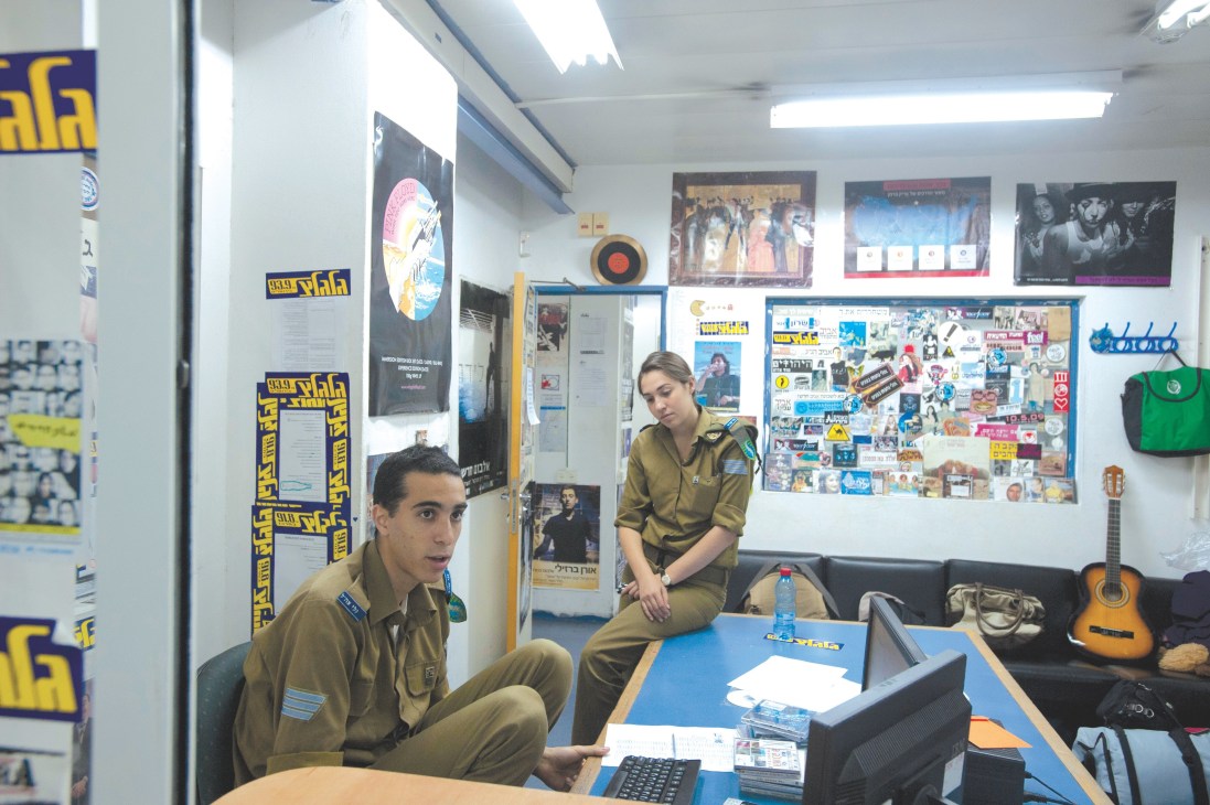 Two Israeli soldiers in khaki uniforms in a radio station office decorated with posters and music memorabilia.
