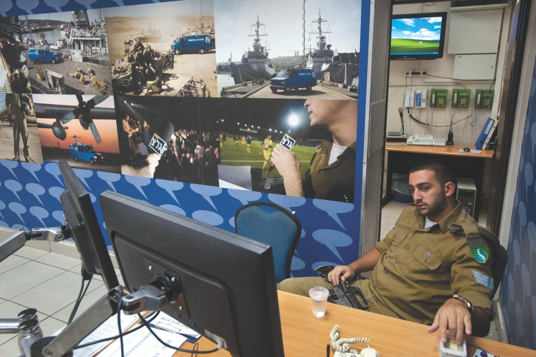 Israeli soldier in khaki uniform sits at a desk in a radio station studio, wearing his rifle.
