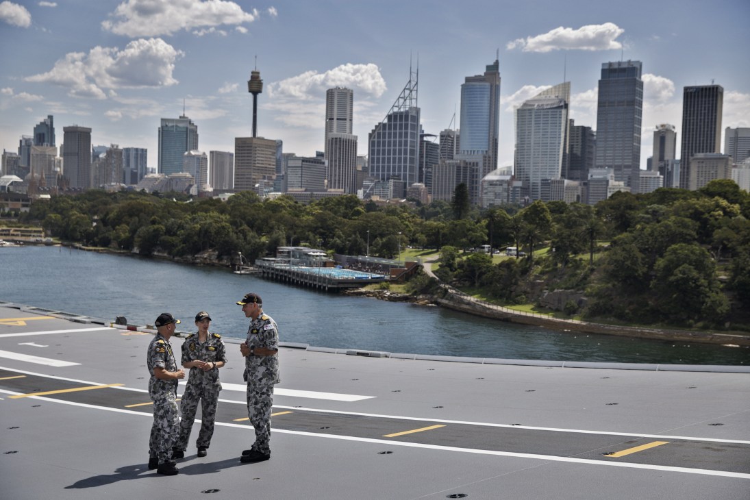 Three naval personnel in camouflage uniforms stand on HMAS Canberra's flight deck overlooking Sydney Harbour and the city skyline.