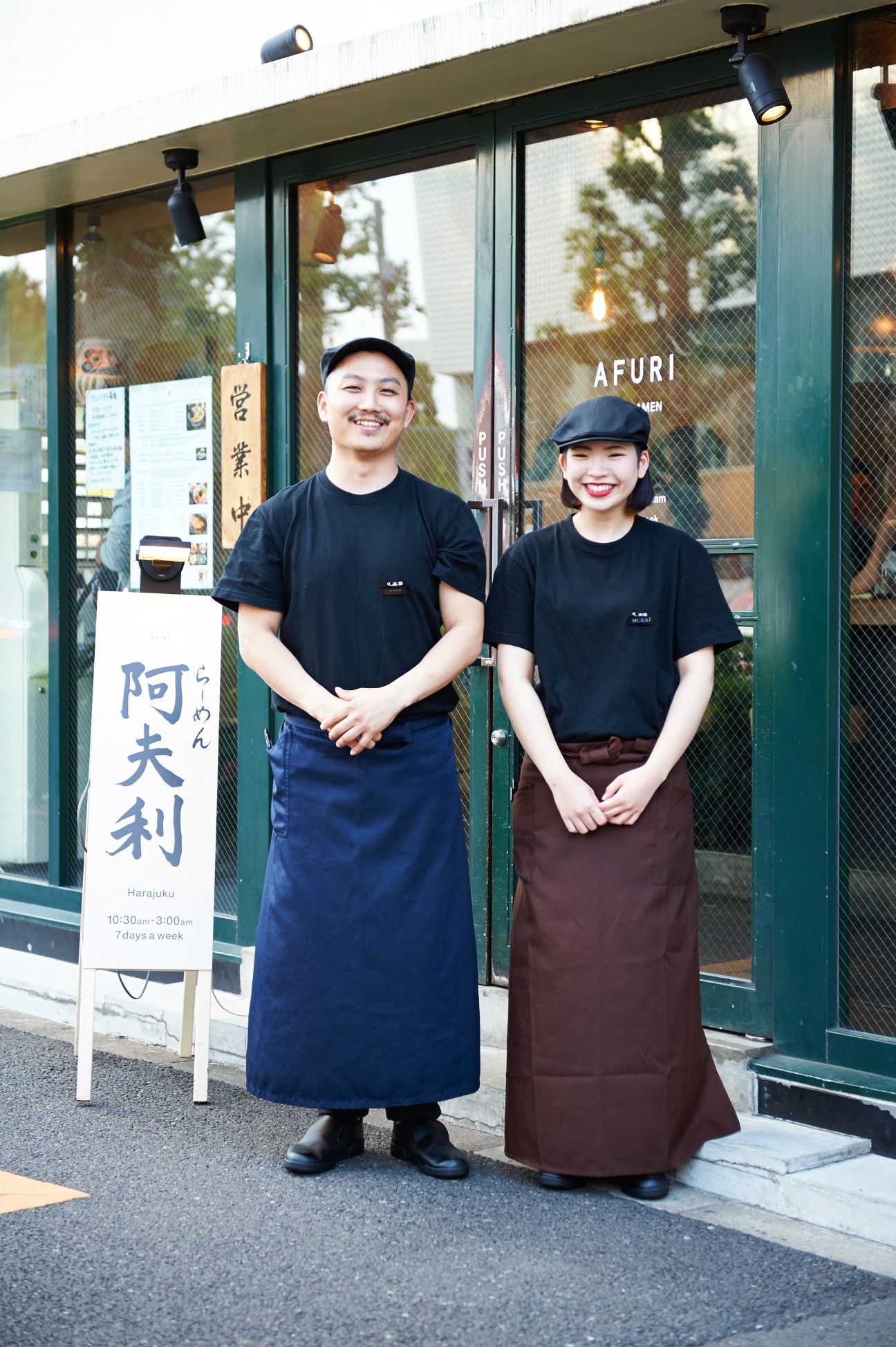Two smiling staff members stand outside Afuri ramen restaurant in Tokyo, wearing black shirts and aprons.